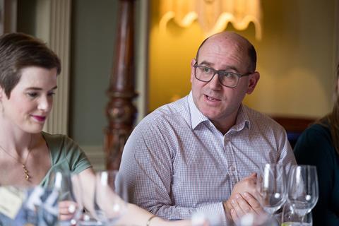 Photography of Allianz fraud manager James Burge engaging in a conversation with a fellow roundtable attendee across the table. Insurance Times editor Katie Scott sits to his right. He wears a brown and blue chequered shirt and brown square glasses.